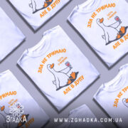 Футболка зла не тримаю, білого кольору, зображена на манекені з блискучою поверхнею, фон світло-сірий, принт з веселою качкою, що тримає келих з вином, текст оранжевого кольору.