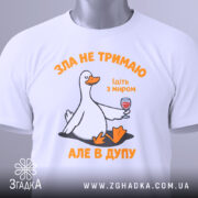 Футболка зла не тримаю, білого кольору, зображена на плоскій поверхні, фон світло-сірий, принт з качкою, що тримає келих з вином, текст оранжевого кольору, на футболці є етикетка з зеленим шнурком.