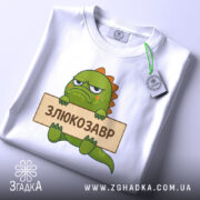 Футболка злюкозавр сердитий, біла, зображена зверху, фон світло-сірий, принт злюкозавра з зеленим тілом, тримає табличку з написом 'ЗЛЮКОЗАВР', на футболці є етикетка з зеленим шнурком.