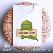 Футболка злюкозавр сердитий, біла, зображена зверху на плетеній кошику, фон білий, принт злюкозавра з зеленим тілом, тримає табличку з написом 'ЗЛЮКОЗАВР'.