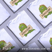 Футболка злюкозавр сердитий, біла, зображена на манекені, фон світло-сірий, принт злюкозавра з зеленим тілом і жовто-оранжевими шипами, тримає табличку з написом 'ЗЛЮКОЗАВР'.