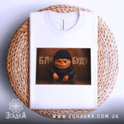 Футболка злий персонаж, біла, складена на плетеній основі, фон нейтральний, принт з злим персонажем у чорній шапці та куртці, текст на фоні.