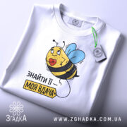 Футболка знайти її вдача, біла, зображена зверху, фон світло-сірий, принт з бджолою та етикеткою, напис 'ЗНАЙТИ ЇЇ – МОЯ ВДАЧА'.