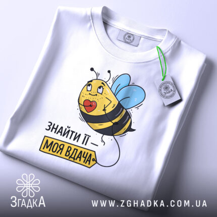 Футболка знайти її вдача, біла, зображена зверху, фон світло-сірий, принт з бджолою та етикеткою, напис 'ЗНАЙТИ ЇЇ – МОЯ ВДАЧА'.