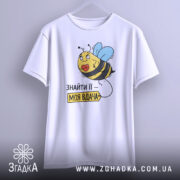 Футболка знайти її вдача, біла, зображена на вішалці, фон світло-сірий, принт з бджолою та написом 'ЗНАЙТИ ЇЇ – МОЯ ВДАЧА'.