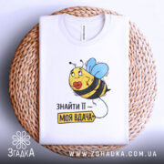 Футболка знайти її вдача, біла, зображена зверху на плетеному кошику, фон світло-сірий, принт з бджолою та написом 'ЗНАЙТИ ЇЇ – МОЯ ВДАЧА'.