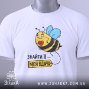 Футболка знайти її вдача, біла, зображена зверху, фон світло-сірий, принт з бджолою та етикеткою, напис 'ЗНАЙТИ ЇЇ – МОЯ ВДАЧА'.