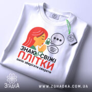 Футболка Знаю свіжі плітки білого кольору, складена на поверхні, фон світло-сірий, принт з дівчиною з червоним волоссям, що шепоче, з елементами замка та діалогових вікон, на комірці є етикетка.