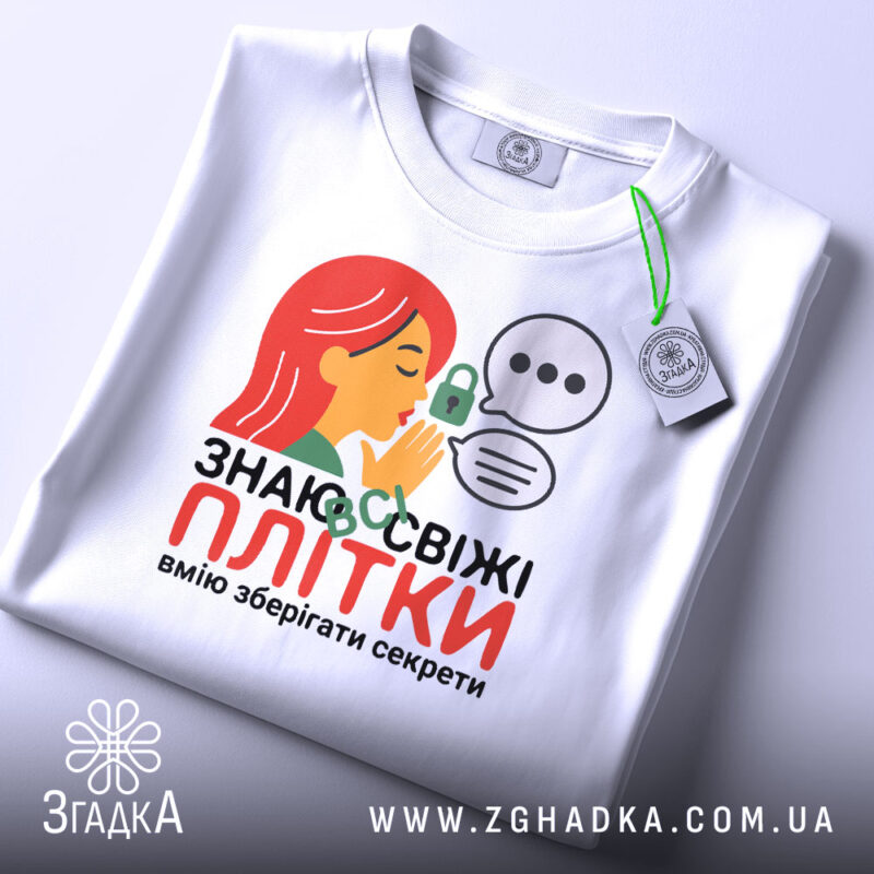 Футболка Знаю свіжі плітки білого кольору, складена на поверхні, фон світло-сірий, принт з дівчиною з червоним волоссям, що шепоче, з елементами замка та діалогових вікон, на комірці є етикетка.