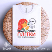 Футболка Знаю свіжі плітки білого кольору, складена на плетеній кошику, фон світлий, принт з дівчиною з червоним волоссям, що шепоче, з елементами замка та діалогових вікон.