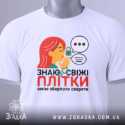 Футболка Знаю свіжі плітки білого кольору, складена на поверхні, фон світло-сірий, принт з дівчиною з червоним волоссям, що шепоче, з елементами замка та діалогових вікон, на комірці є етикетка.