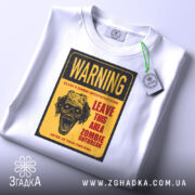 Футболка зомбі попередження, біла, складена на поверхні, фон світлий, принт з черепом та текстом жовтого кольору, з етикеткою.