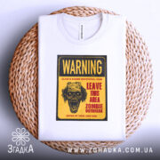 Футболка зомбі попередження, біла, складена на плетеній кошику, фон нейтральний, принт з черепом та текстом жовтого кольору.