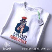 Футболка zombie attack білого кольору, складена на поверхні, фон світло-сірий, принт з черепом у капелюсі зірки та написом 'ARE YOU READY FOR A ZOMBIE ATTACK' у червоному та синьому кольорах, з етикеткою на рукаві.