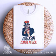Футболка zombie attack білого кольору, складена на плетеній кошику, фон білий, принт з черепом у капелюсі зірки та написом 'ARE YOU READY FOR A ZOMBIE ATTACK' у червоному та синьому кольорах.