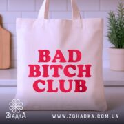 Шопер Bad Bitch Club на кухонному фоні, бежевого кольору, з фронтальним ракурсом, товщина тканини середня, ручки короткі, місткість велика, принт з написом 'BAD BITCH CLUB' червоного кольору.
