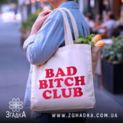 Шопер Bad Bitch Club на вулиці, бежевого кольору, з ракурсом ззаду, товщина тканини середня, ручки довгі, місткість велика, принт з написом 'BAD BITCH CLUB' червоного кольору, наповнений продуктами.
