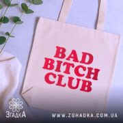 Шопер Bad Bitch Club на фоні з рослинами, бежевого кольору, з ракурсом зверху, товщина тканини середня, ручки середньої довжини, місткість велика, принт з написом 'BAD BITCH CLUB' червоного кольору.