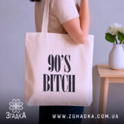 Шопер для дитини 90-х, бежевий, зображений з боку, на фоні кімнати з рослинами, тканина середньої товщини, ручки середньої довжини, місткість підходить для шопінгу, принт з написом '90'S BITCH' великими літерами.