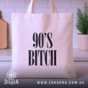 Шопер для дитини 90-х, бежевий, зображений з фронтального ракурсу на фоні кухонних предметів, тканина середньої товщини, ручки середньої довжини, місткість достатня для продуктів, принт з написом '90'S BITCH' великими літерами.