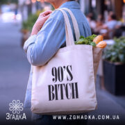 Шопер для дитини 90-х, бежевий, зображений з боку на фоні вулиці, тканина середньої товщини, ручки середньої довжини, місткість достатня для покупок, принт з написом '90'S BITCH' великими літерами.