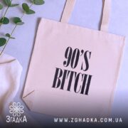 Шопер для дитини 90-х, бежевий, зображений зверху на фоні текстильних виробів та рослин, тканина середньої товщини, ручки середньої довжини, місткість підходить для повсякденних речей, принт з написом '90'S BITCH' великими літерами.