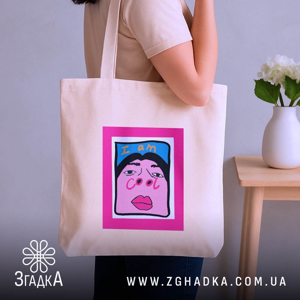 Шопер для класних бежевий, з ракурсом збоку, на фоні нейтральної стіни, з тонкої бавовняної тканини, довжина ручок середня, місткість достатня для покупок, принт з написом 'I am cool' на яскравому рожевому фоні.