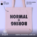 Шопер для НЕнормальних. Бежевий, знімок з фронтального ракурсу на білому фоні, тканина середньої товщини, ручки середньої довжини, місткість достатня для повсякденних речей, принт чорного кольору з написом 'NORMAL is BORING'.