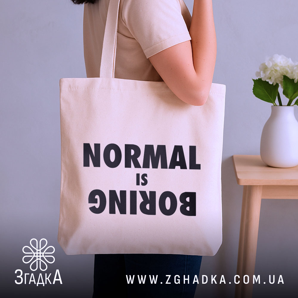 Шопер для НЕнормальних. Бежевий, знімок з боку, на фоні кімнати з рослинами, тканина середньої товщини, ручки середньої довжини, місткість достатня для повсякденних речей, принт чорного кольору з написом 'NORMAL is BORING'.