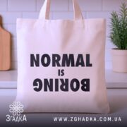 Шопер для НЕнормальних. Бежевий, знімок з фронтального ракурсу на кухонному фоні, тканина середньої товщини, ручки середньої довжини, місткість достатня для повсякденних речей, принт чорного кольору з написом 'NORMAL is BORING'.