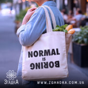 Шопер для НЕнормальних. Бежевий, знімок з боку на фоні міського пейзажу, тканина середньої товщини, ручки середньої довжини, місткість достатня для продуктів, принт чорного кольору з написом 'NORMAL is BORING'.