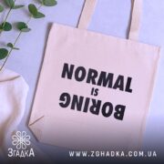 Шопер для НЕнормальних. Бежевий, знімок зверху на фоні з рослинами, тканина середньої товщини, ручки середньої довжини, місткість достатня для повсякденних речей, принт чорного кольору з написом 'NORMAL is BORING'.
