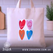 Шопер для закоханих. Бежевий, зображення спереду, на кухонному фоні. Тканина середньої товщини, ручки середньої довжини, місткість достатня для повсякденних речей, принт з серцями та написом 'LOVE' у яскравих кольорах.