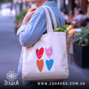 Шопер для закоханих. Бежевий, зображення спереду, на вулиці з розмитим фоном. Тканина середньої товщини, ручки середньої довжини, місткість достатня для продуктів, принт з серцями та написом 'LOVE' у яскравих кольорах.