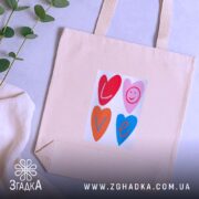 Шопер для закоханих. Бежевий, зображення спереду, на нейтральному фоні з рослинами. Тканина середньої товщини, ручки середньої довжини, місткість достатня для повсякденних речей, принт з серцями та написом 'LOVE' у яскравих кольорах.