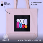 Шопер Good Mood, бежевого кольору, зображений з близької відстані на світлому фоні, з товстої бавовняної тканини, ручки середньої довжини, місткий, з принтом на чорному фоні з написом 'GOOD MOOD' різнокольоровими літерами.