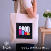 Шопер Good Mood, бежевого кольору, показаний на фоні з сірою стіною, з товстої бавовняної тканини, ручки середньої довжини, місткий, з принтом на чорному фоні з написом 'GOOD MOOD' різнокольоровими літерами, на плечі людини.