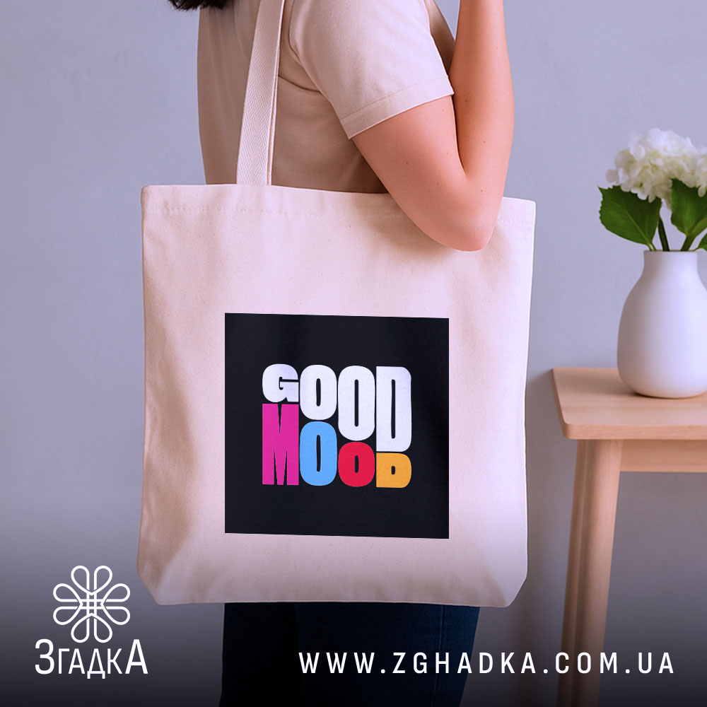 Шопер Good Mood, бежевого кольору, показаний на фоні з сірою стіною, з товстої бавовняної тканини, ручки середньої довжини, місткий, з принтом на чорному фоні з написом 'GOOD MOOD' різнокольоровими літерами, на плечі людини.
