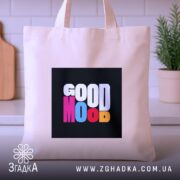 Шопер Good Mood, бежевого кольору, зображений з близької відстані на світлому фоні кухні, з товстої бавовняної тканини, ручки середньої довжини, місткий, з принтом на чорному фоні з написом 'GOOD MOOD' різнокольоровими літерами.