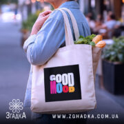 Шопер Good Mood, бежевого кольору, показаний з боку на фоні вулиці, з товстої бавовняної тканини, ручки середньої довжини, місткий, з принтом на чорному фоні з написом 'GOOD MOOD' різнокольоровими літерами, в руках людини з продуктами.