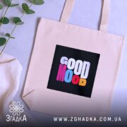 Шопер Good Mood, бежевого кольору, зображений на фоні з рослинами, з товстої бавовняної тканини, ручки середньої довжини, місткий, з принтом на чорному фоні з написом 'GOOD MOOD' різнокольоровими літерами.