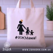 Шопер яжмама, бежевий, з фронтальної сторони, на кухонному фоні, тонка бавовняна тканина, короткі ручки, місткий, принт з жінкою в сукні та двома дітьми, напис #ЯЖМама!