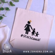 Шопер яжмама, бежевий, з фронтальної сторони, на нейтральному фоні з рослинами, тонка бавовняна тканина, короткі ручки, місткий, принт з жінкою в сукні та двома дітьми, напис #ЯЖМама!