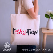 Шопер любовна революція, бежевого кольору, зображений з боку людини, на фоні кімнати з рослинами, виготовлений з тонкої бавовняної тканини, ручки середньої довжини, місткий, з принтом 'revoluTion' у червоному та чорному кольорах.