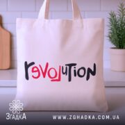 Шопер любовна революція, бежевого кольору, зображений з фронтальної сторони, на фоні кухонного інтер'єру, виготовлений з тонкої бавовняної тканини, ручки середньої довжини, місткий, з принтом 'revoluTion' у червоному та чорному кольорах.