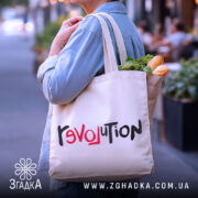 Шопер любовна революція, бежевого кольору, зображений з боку людини, на фоні вулиці, виготовлений з тонкої бавовняної тканини, ручки середньої довжини, місткий, з принтом 'revoluTion' у червоному та чорному кольорах.