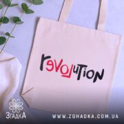 Шопер любовна революція, бежевого кольору, зображений зверху, на фоні світлого столу з рослинами, виготовлений з тонкої бавовняної тканини, ручки середньої довжини, місткий, з принтом 'revoluTion' у червоному та чорному кольорах.