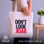 Шопер не дивись назад. Бежевий колір, ракурс з боку, фон нейтральний. Тканина середньої товщини, ручки довгі. Місткість велика, принт з написом 'DON'T LOOK BACK' в чорному та червоному кольорах.