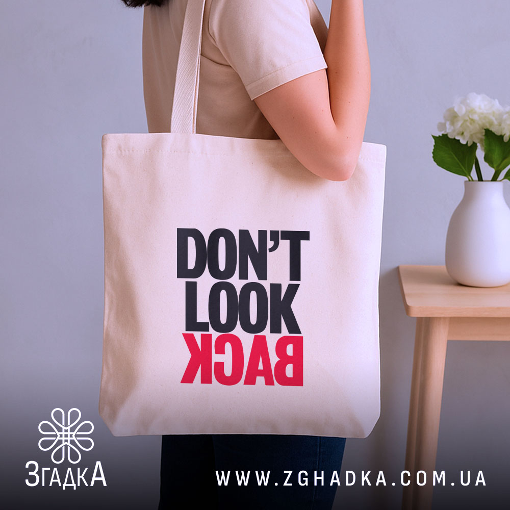 Шопер не дивись назад. Бежевий колір, ракурс з боку, фон нейтральний. Тканина середньої товщини, ручки довгі. Місткість велика, принт з написом 'DON'T LOOK BACK' в чорному та червоному кольорах.