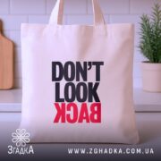 Шопер не дивись назад. Бежевий колір, фронтальний ракурс, фон кухонний. Тканина середньої товщини, ручки короткі. Місткість середня, принт з написом 'DON'T LOOK BACK' в чорному та червоному кольорах.